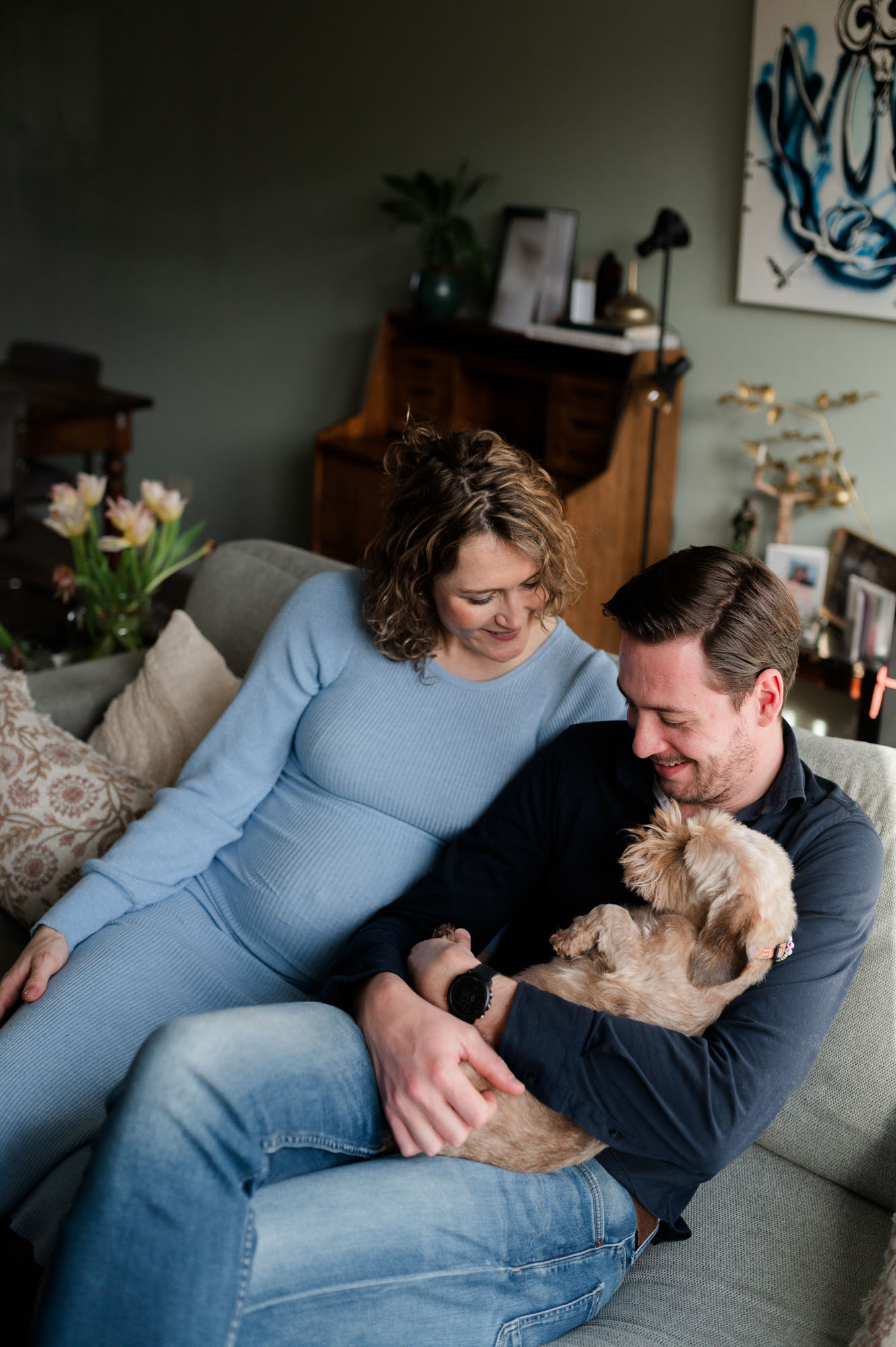 Zwangere vrouw met partner en hond, genietend en lachend op de bank tijdens een fotoshoot.