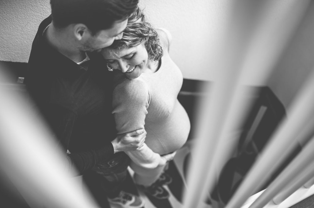Zwanger koppel knuffelt, gefotografeerd door de spijlen van de trap.
