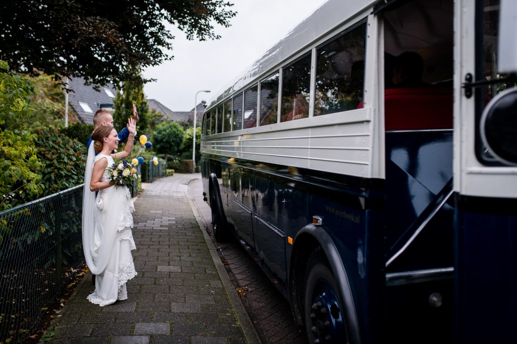 Bruidspaar die hun gasten glimlachend toe zwaait. De daggasten rijden in een grote bus naar de bruiloft ceremonie.